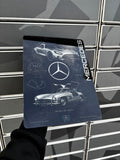 A person holds a rectangular metal mouse pad featuring Mercedes-Benz branding and classic car designs. The mouse pad is held in front of a gray tiled wall.