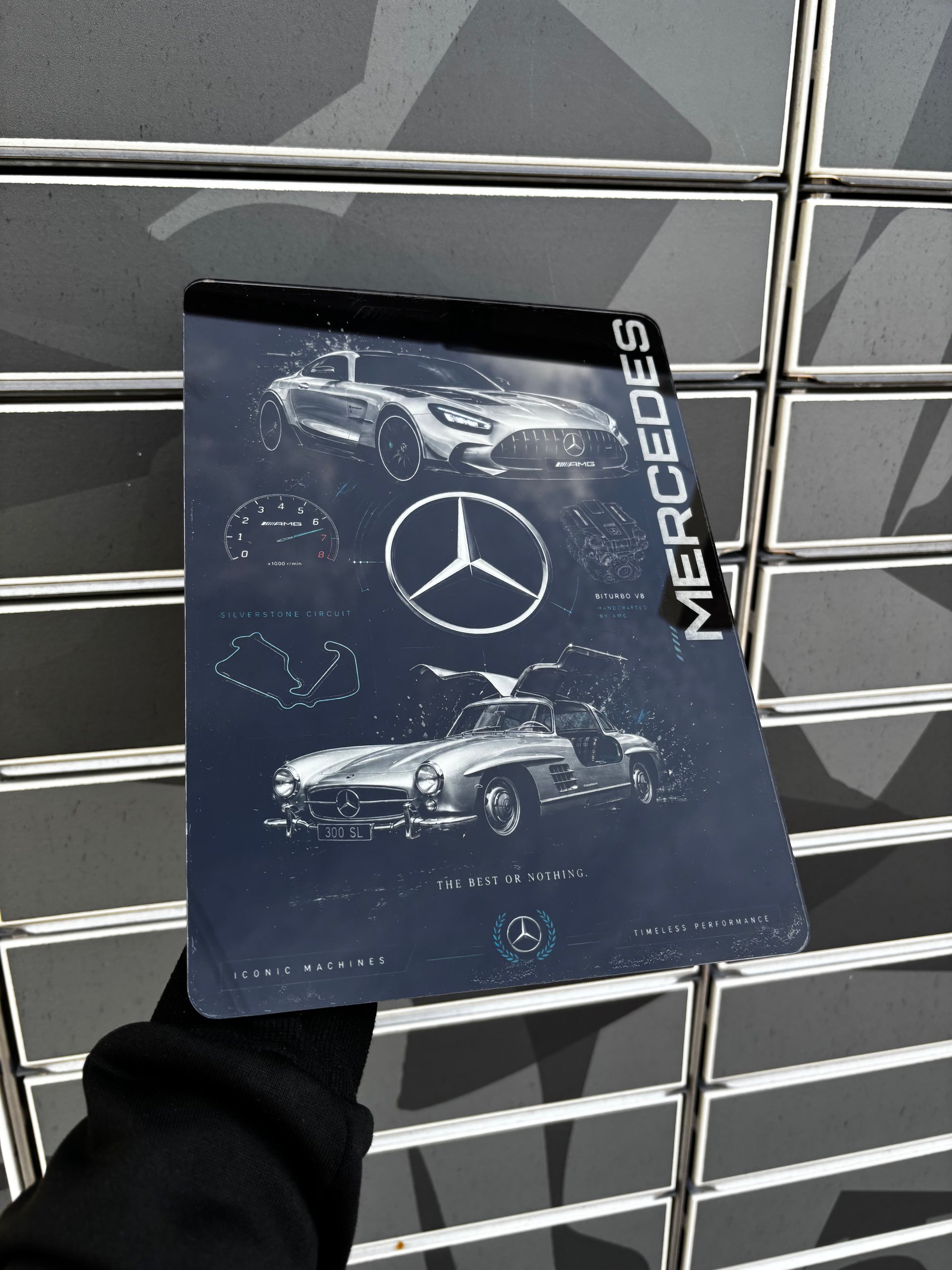 A person holds a rectangular metal mouse pad featuring Mercedes-Benz branding and classic car designs. The mouse pad is held in front of a gray tiled wall.