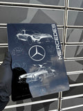 A hand wearing a black glove holds a rectangular metal print featuring Mercedes-Benz branding, a silver sports car illustration, and technical-style graphics, in front of a gray metal grid background.