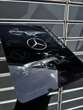 A glossy rectangular Mercedes-Benz branded plaque or sign featuring stylized car illustrations and the Mercedes logo, photographed against a gray tiled wall.