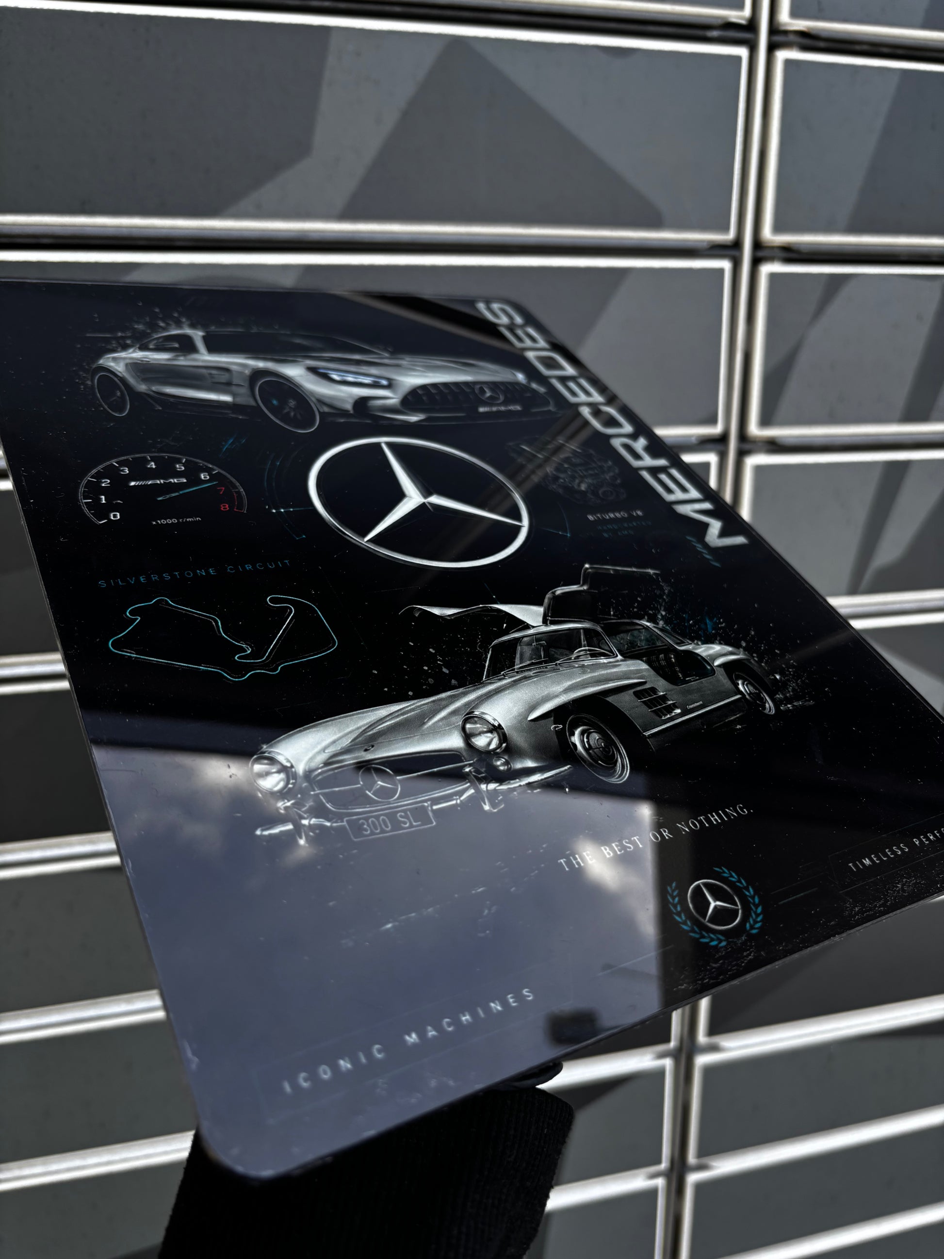 A glossy rectangular Mercedes-Benz branded plaque or sign featuring stylized car illustrations and the Mercedes logo, photographed against a gray tiled wall.