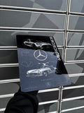A person's hand in a black sleeve holds a glossy Mercedes-Benz Leaning Machine book against a gray slatted metal wall.