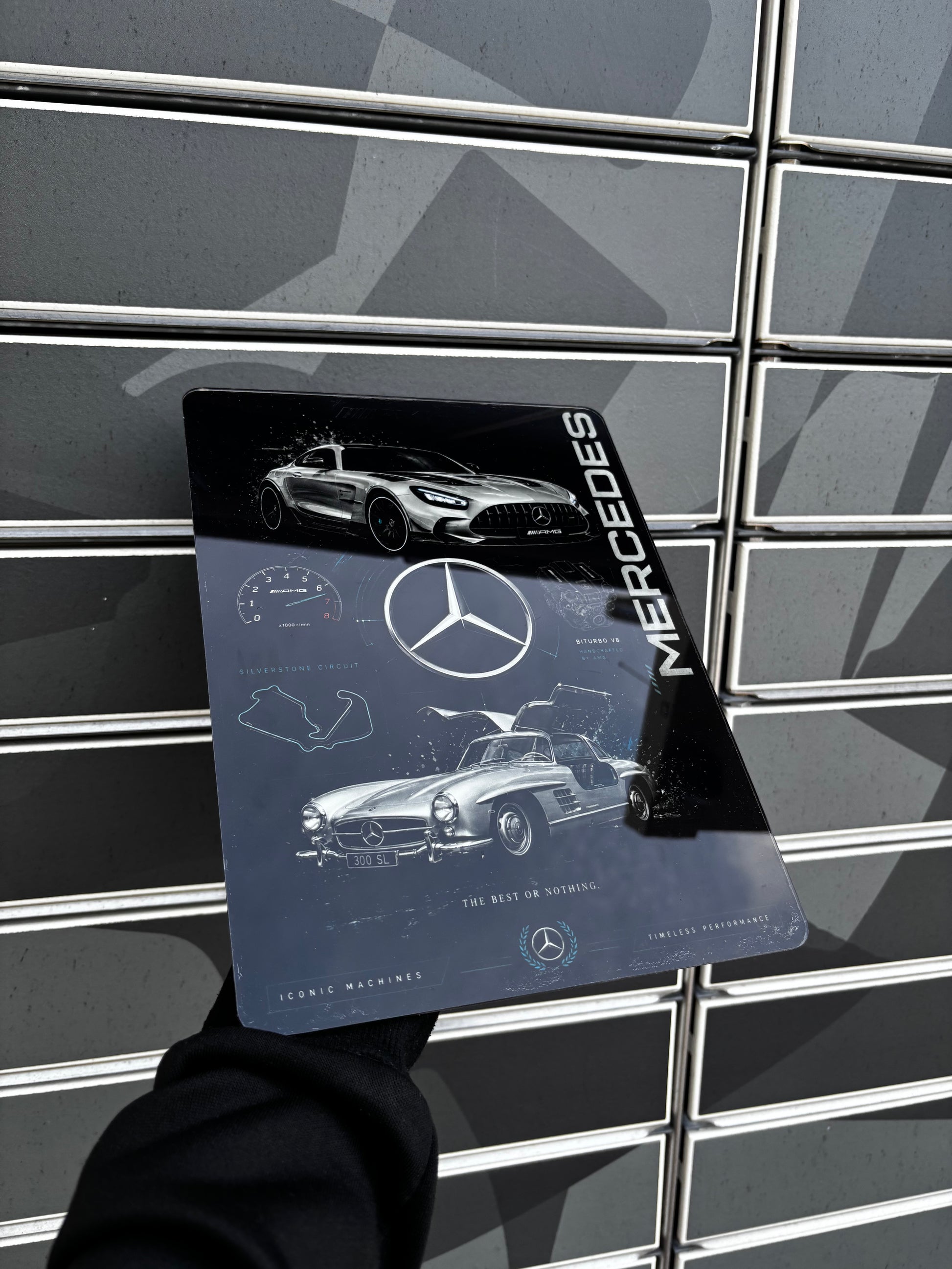 A person's hand in a black sleeve holds a glossy Mercedes-Benz Leaning Machine book against a gray slatted metal wall.