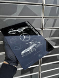 A hand wearing a black glove holds a glossy Mercedes-Benz themed mousepad or flat printed card featuring white car illustrations, the Mercedes logo, and the word MERCEDES on a dark background, set against a gray slatted wall.